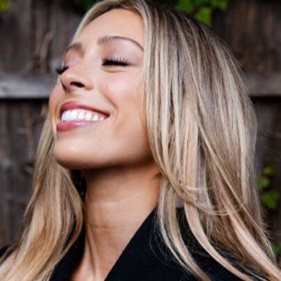Young singer Jemma Cher smiling with her face turned slightly upwards, eyes closed, with long blonde hair, posing outdoors in front of a fence.