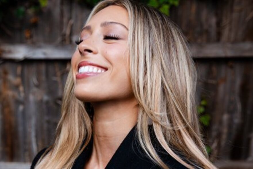 Young singer Jemma Cher smiling with her face turned slightly upwards, eyes closed, with long blonde hair, posing outdoors in front of a fence.