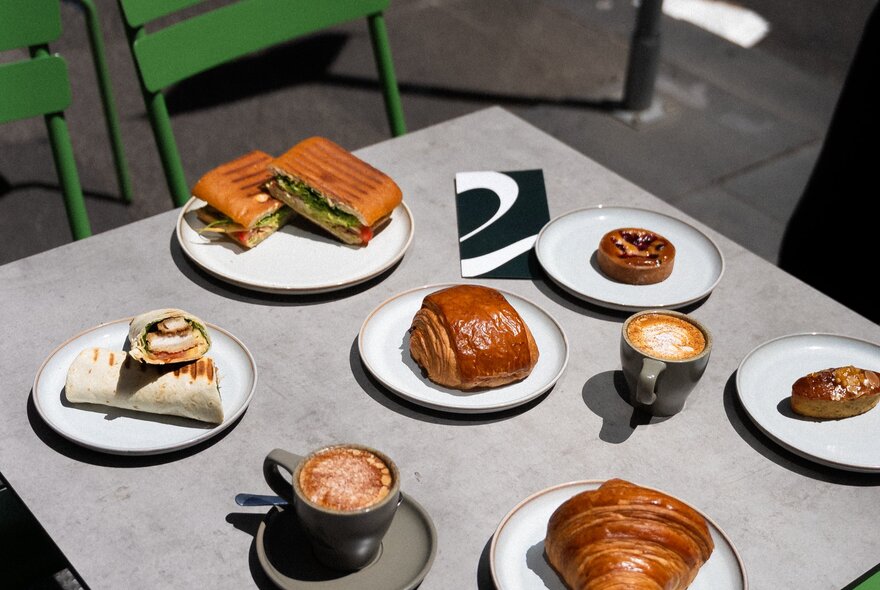 A cafe table with many small plates of pastries and toasties with two cups of coffee.