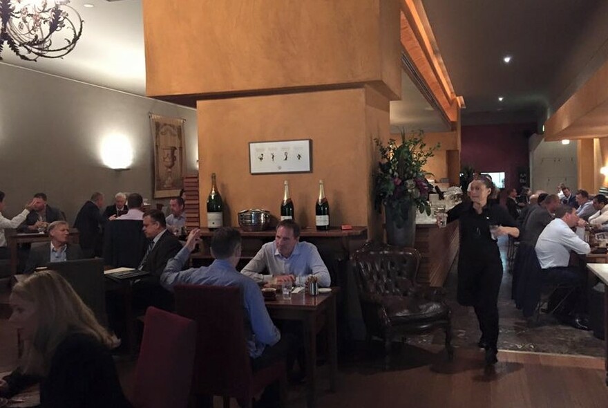 Cafe interior with people seated at tables, wooden feature wall, chandelier and bottles of bubbles.