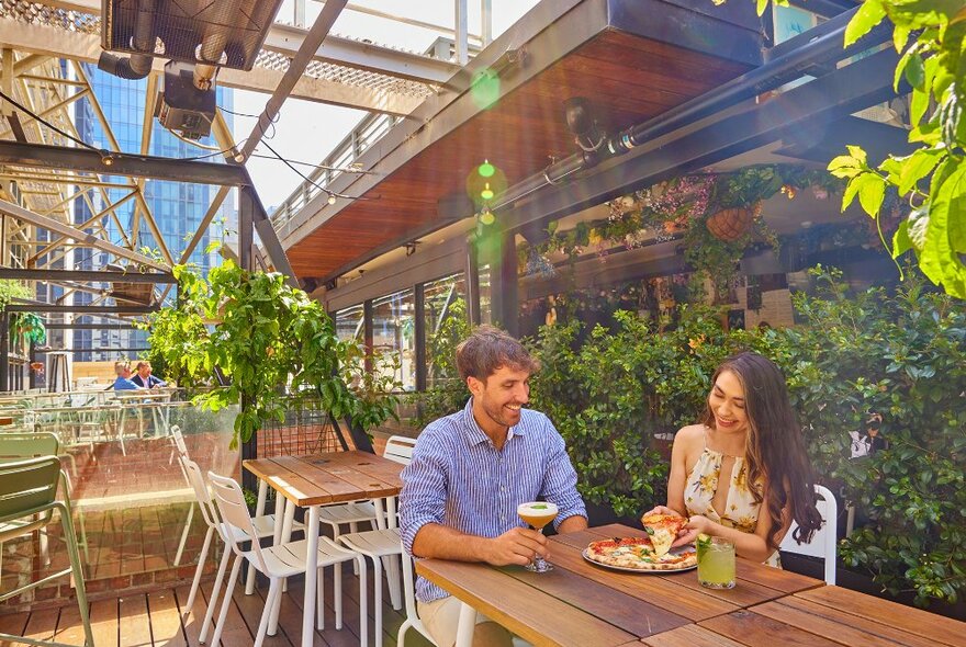 A couple sharing a pizza and cocktails on a sunny rooftop. 