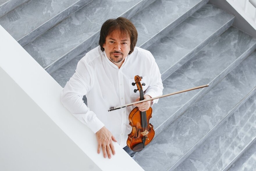 Musician holding a violin, standing on steps and looking up, dressed in white.