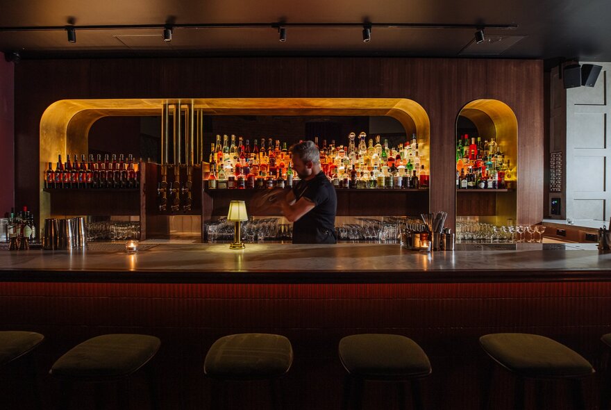 The dimly lit central bar of Death & Co, with high bar stools, a bartender behind the curved bar, and bottles lined up on the back wall.