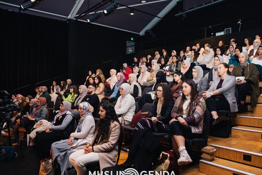 Audience members in a seated auditorium with black walls and roof.