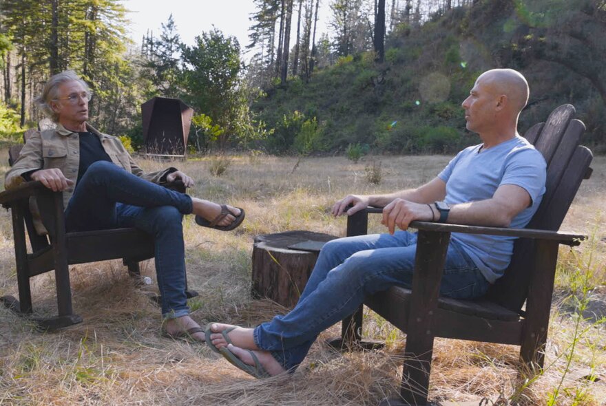 Two middle-aged men seated outdoors in a field in large wooden chairs, talking to each other.