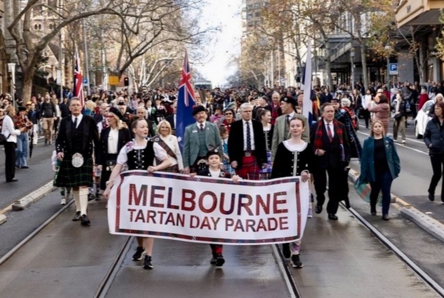 A parade of people walking down Collins Street behind a banner that reads 'MELBOURNE TARTAN DAY PARADE'