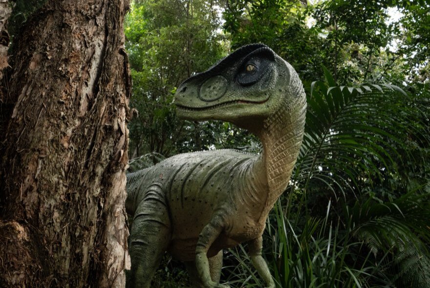 A large dinosaur model beside a tree trunk in a park setting. 