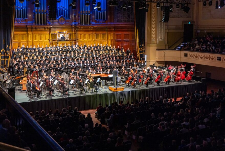 A large group of orchestra musicians and choir singers on a stage, a grand organ behind them, and people in seats listening to the concert being performed.