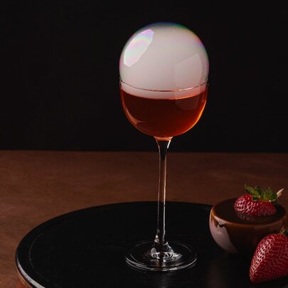 A red cocktail with a cloudy dome, on a black tray next to two strawberries.