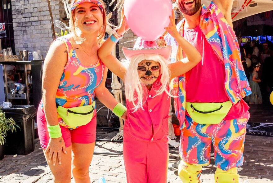 Family wearing day-glo shorts, child in a pink onesie with blond wig, face makeup and cowboy hat holding a balloon.