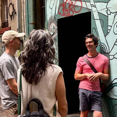 A tour guide wearing a pink t-shirt, talking to a couple on a walking tour about the mural art on the wall behind him.