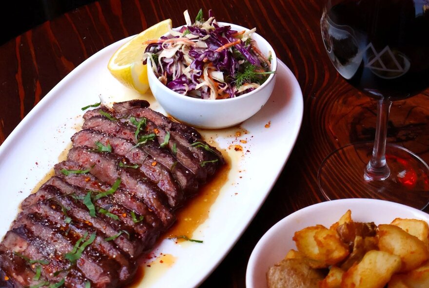 A plate of sliced steak with salad, potatoes and red wine.
