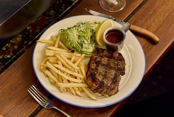 A steak on a plate covered in a sauce with fries, salad and a small ramekin of tomato sauce.