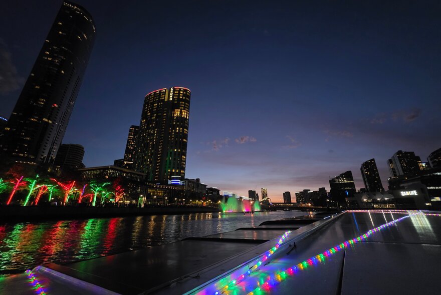 Bright lights and water fountains near the edge of the Yarra River, with the city skyline in the background, as seen from a boat cruise at night.