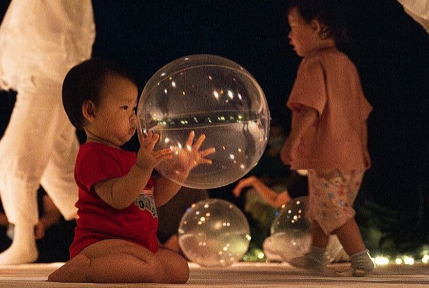 A baby playing with a large bubble, other children and figures in the background.