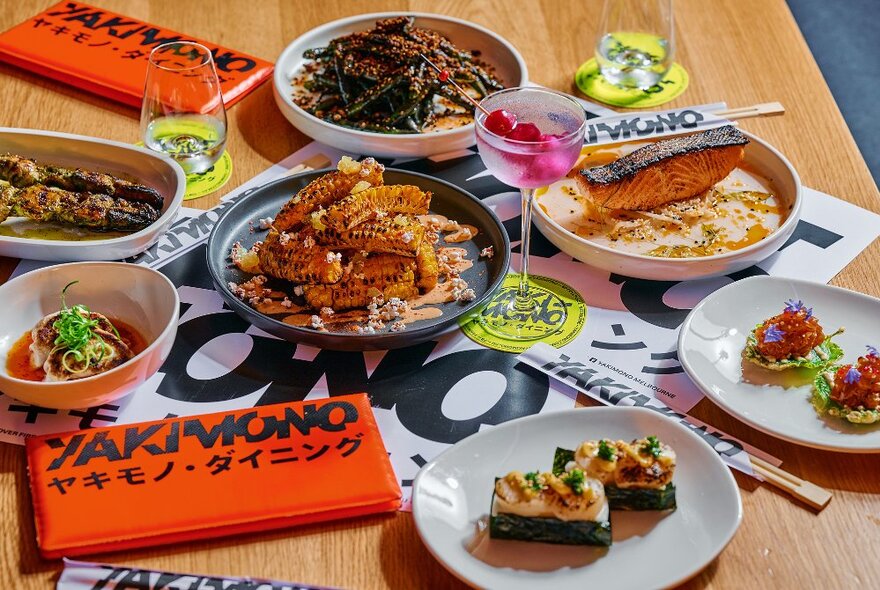 Several plates of Japanese food arranged on a restaurant dining table, with a pink cocktail and menus.