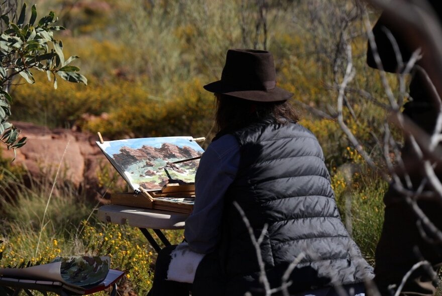Person painting en plein air amongst grasses and foliage.