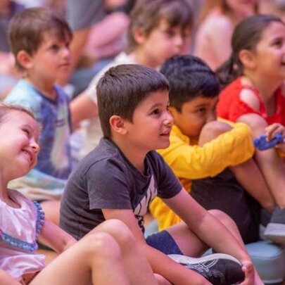 Children sitting cross legged on the floor looking up at someone out of frame.