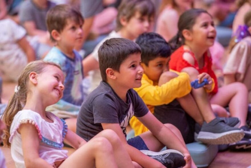 Children sitting cross legged on the floor looking up at someone out of frame.
