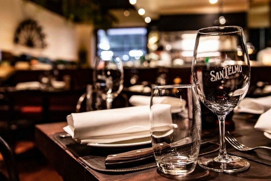 Wine glasses, napkin and cutlery on wooden table inside a restaurant.