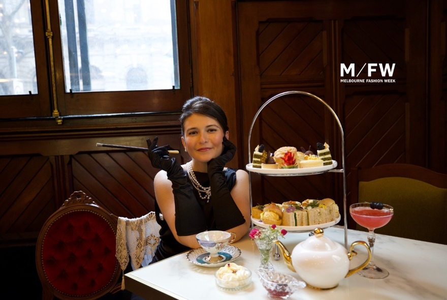 Woman wearing an Audrey Hepburn-inspired little black dress with gloves and jewellery, seated at a table with tiered sandwich and cake stand, tea and cocktail.