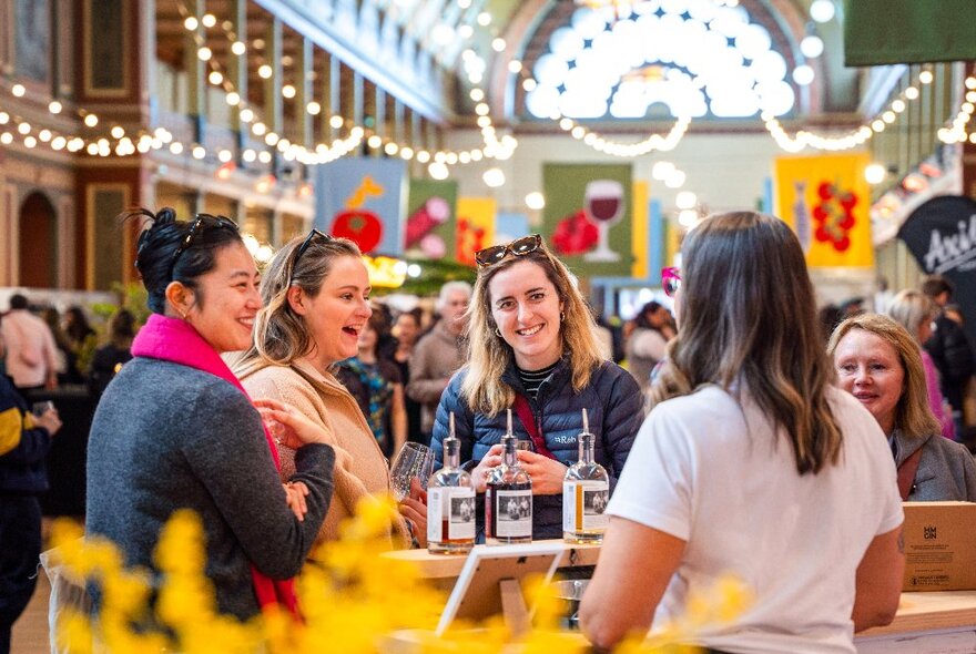 Attendees at The Little Food Market at the Royal Exhibition Building, interacting with a smiling stall holder and being handed some drinks to sample; twinkling fairy lights decorating the large space behind them.