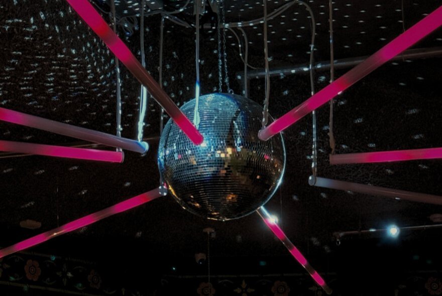 A mirror disco ball against a black twinkling ceiling, with hanging horizontal pink neon glow sticks surrounding it.
