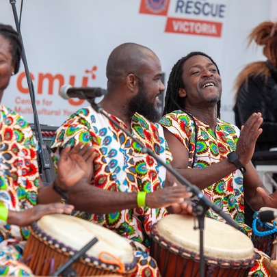 Three African drummers wearing colourful clothing while drumming with their hands on stage.