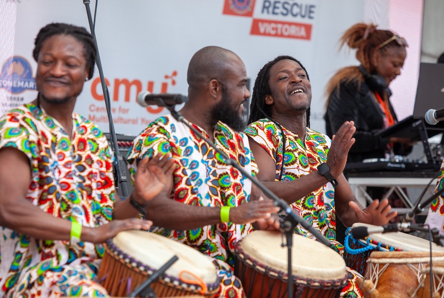 Three African drummers wearing colourful clothing while drumming with their hands on stage.