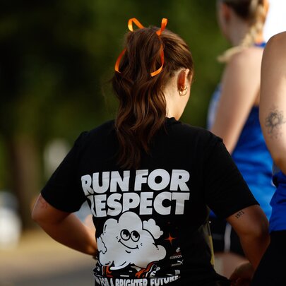 Close-up shot from behind showing a person with long brown hair tied up wearing a black t-shirt that reads 'RUN FOR RESPECT' in bold white lettering, with blurred people in the background wearing race bibs associated with fun runs.