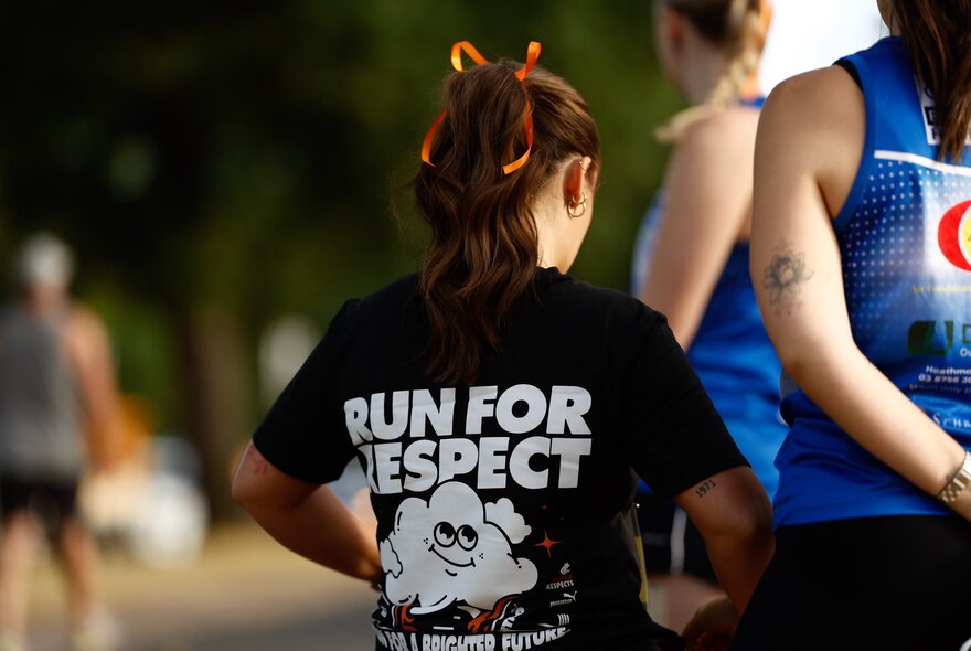 Close-up shot from behind showing a person with long brown hair tied up wearing a black t-shirt that reads 'RUN FOR RESPECT' in bold white lettering, with blurred people in the background wearing race bibs associated with fun runs.