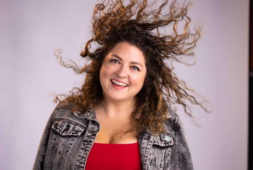 Performer smiling with her hair swirling around her head, wearing a denim jacket and red top.