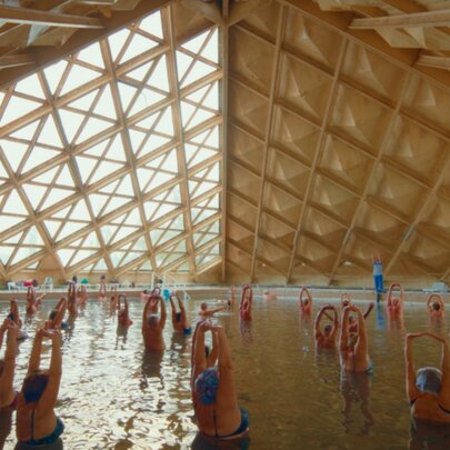 Women bathing and stretching their arms above their heads in a Soviet indoor salt pool underneath a geometric pyramid-shaped roof structure.