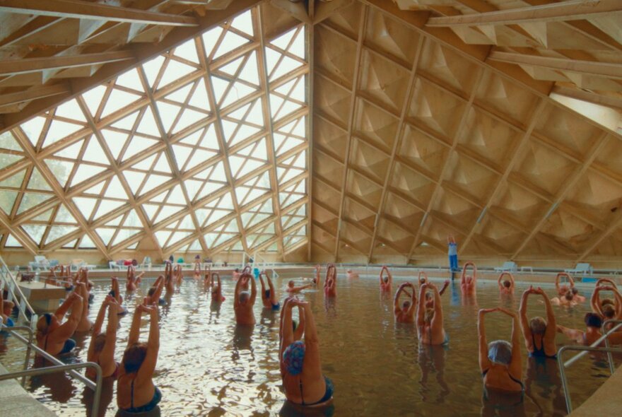 Women bathing and stretching their arms above their heads in a Soviet indoor salt pool underneath a geometric pyramid-shaped roof structure.