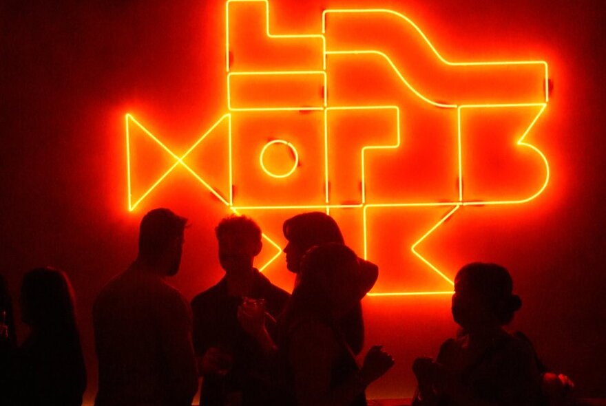 Silhouettes of people at a bar in front of a red-orange neon wall light. 