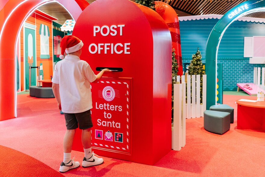 A boy wearing a santa hat posting a letter to Santa at a large red post box in Myer's Santaland.