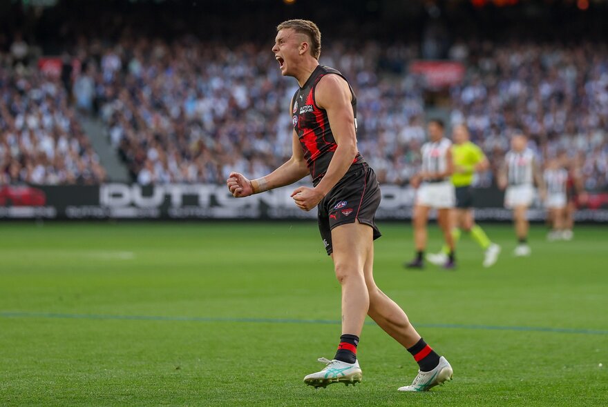An Essendon AFL player celebrating on the field with two clenched fists, blurred players and footy fans in the background.