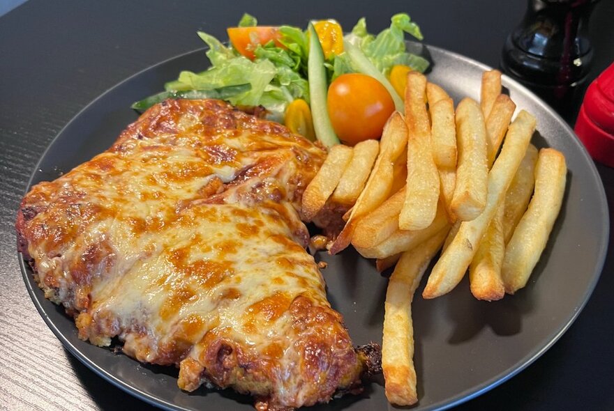 Cheese-covered chicken parmagiana with chips and garden salad on a black plate.