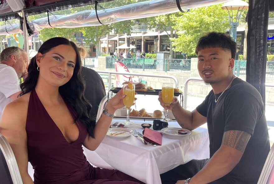 A couple raising their glasses at a dining table on a open-sided boat. 
