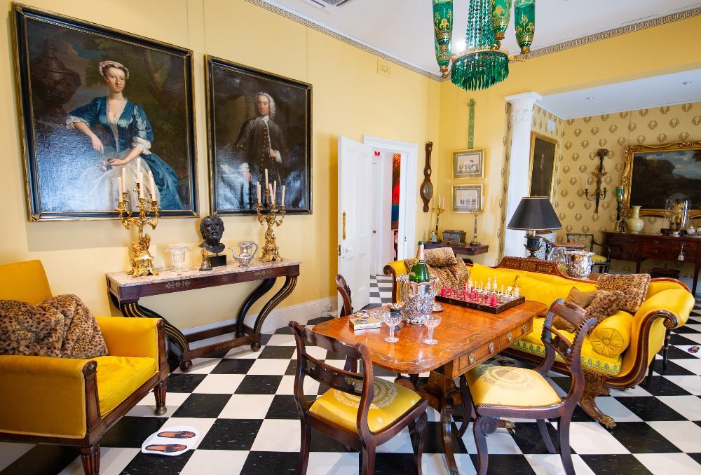 A yellow room inside a historic house with antique furniture there are two paintings of people on the wall.