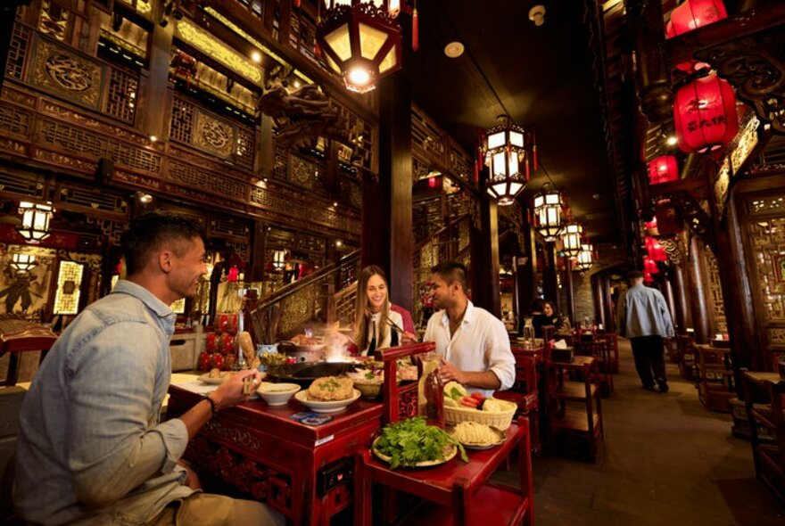 Three people sat at a red table eating hotpot and talking. 