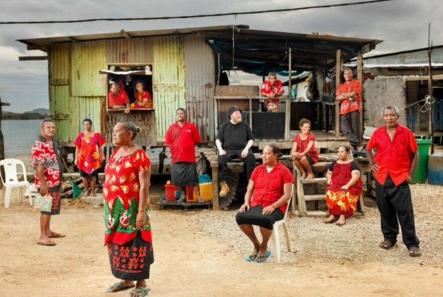 Members of Papua New Guinea’s Tatana Village choir wearing red and black clothes and either standing or seated in front of a makeshift dwelling made of corrugated sheets of metal and wood.