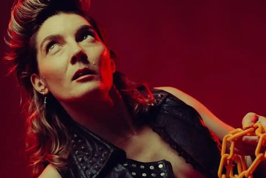 A woman reclining and looking up, wearing a studded leather vest and holding an orange chain.