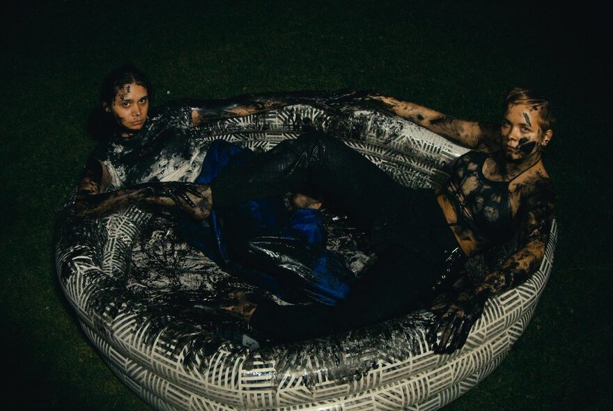 Two people reclined in a small inflatable empty wading pool, both of them smeared in black paint with black paint covering the sides and base of the pool.