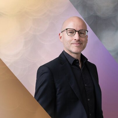 Pianist Daniel de Borah, a young bald man, smiling slightly, he wears glasses and a dark shirt and jacket. 