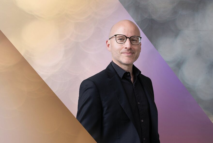 Pianist Daniel de Borah, a young bald man, smiling slightly, he wears glasses and a dark shirt and jacket. 