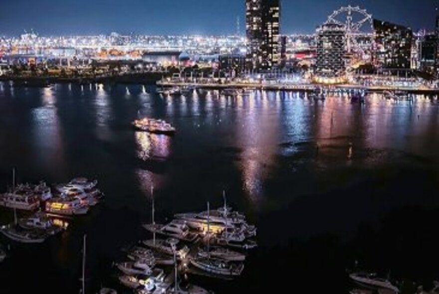 Docklands at night with buildings, boats and wheel lit up and reflected in the water.