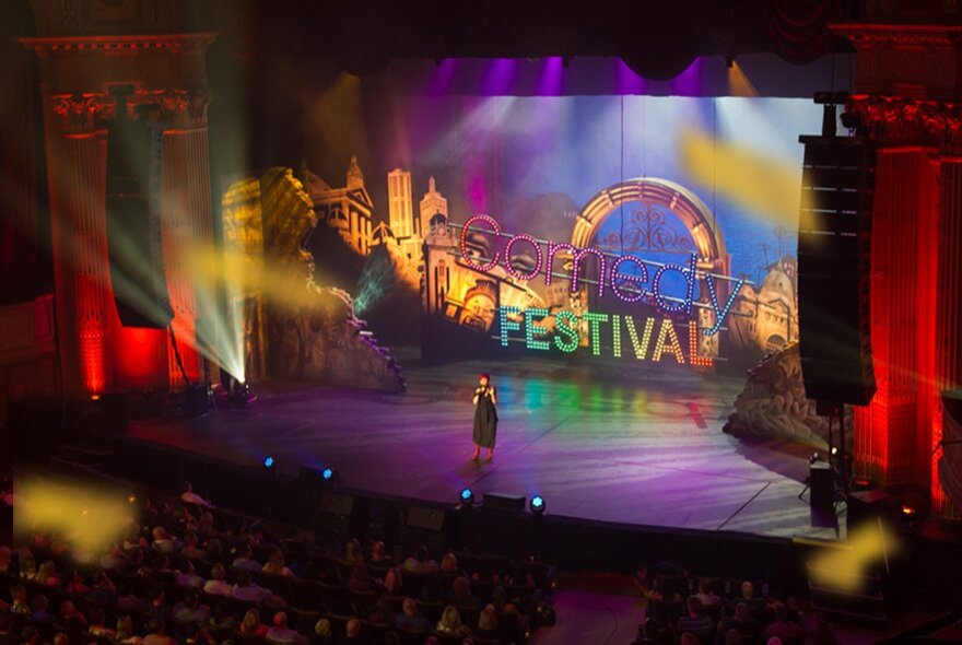 A lone comedian on a huge stage in front of an audience, the backdrop is colourful and has the word FESTIVAL across an image of Melbourne. 