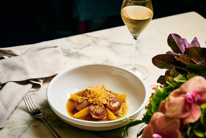 A shallow white bowl of food, on a restaurant table, with a glass of wine, fork and cloth napkin.