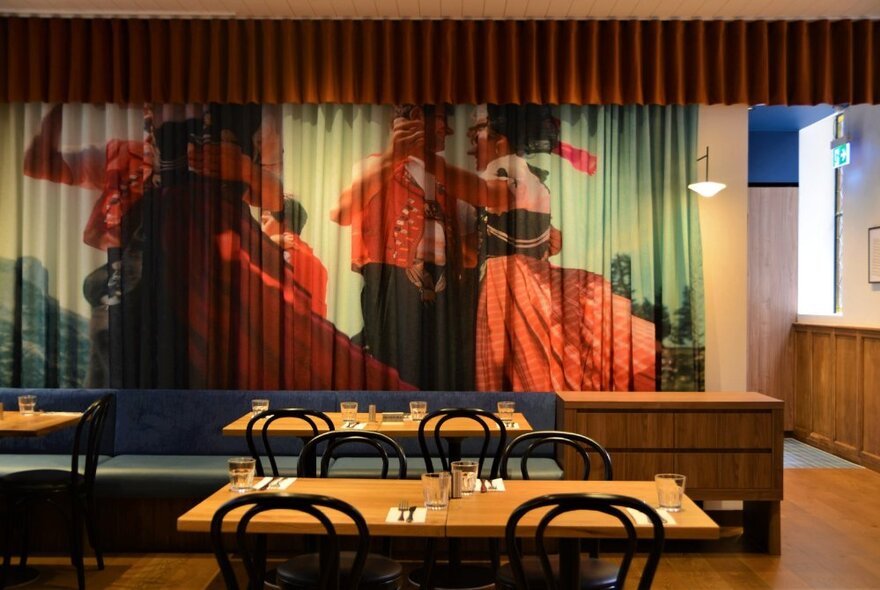 Cafe tables and chairs in an uncluttered restaurant space with wooden details and curtain mural of people dancing.